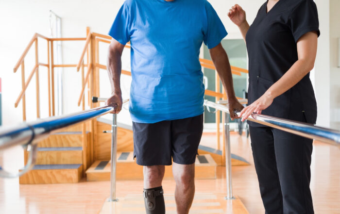 Man wearing a blue tshirt and black gym shorts walking in a physical therapy clinic between the parallel bars, wearing sneakers on both feet and an AFO Sstep AFO Sock, female physical therapist standing on the right side of the parallel bars