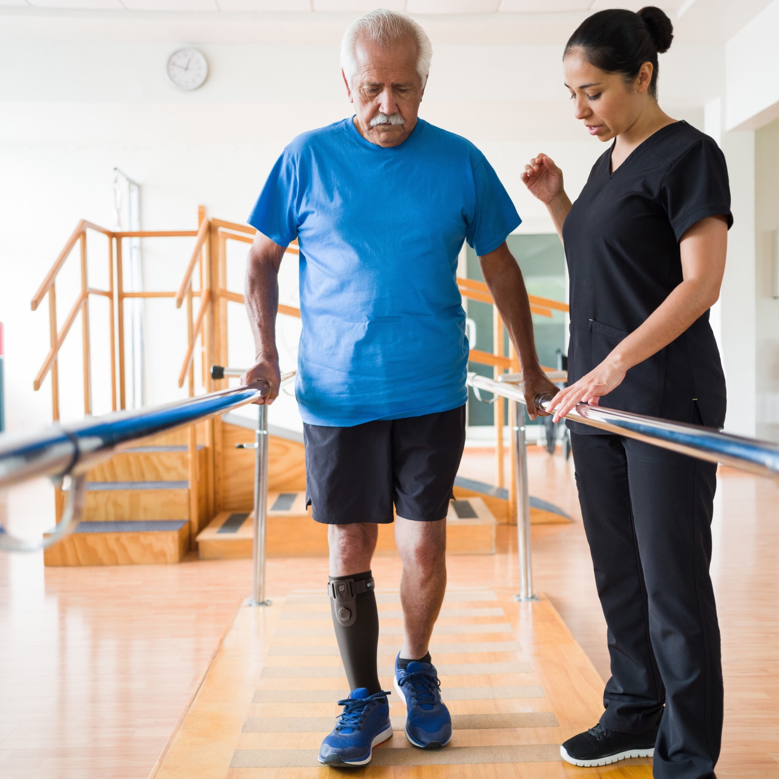 Man wearing a blue tshirt and black gym shorts walking in a physical therapy clinic between the parallel bars, wearing sneakers on both feet and an AFO Sstep AFO Sock, female physical therapist standing on the right side of the parallel bars